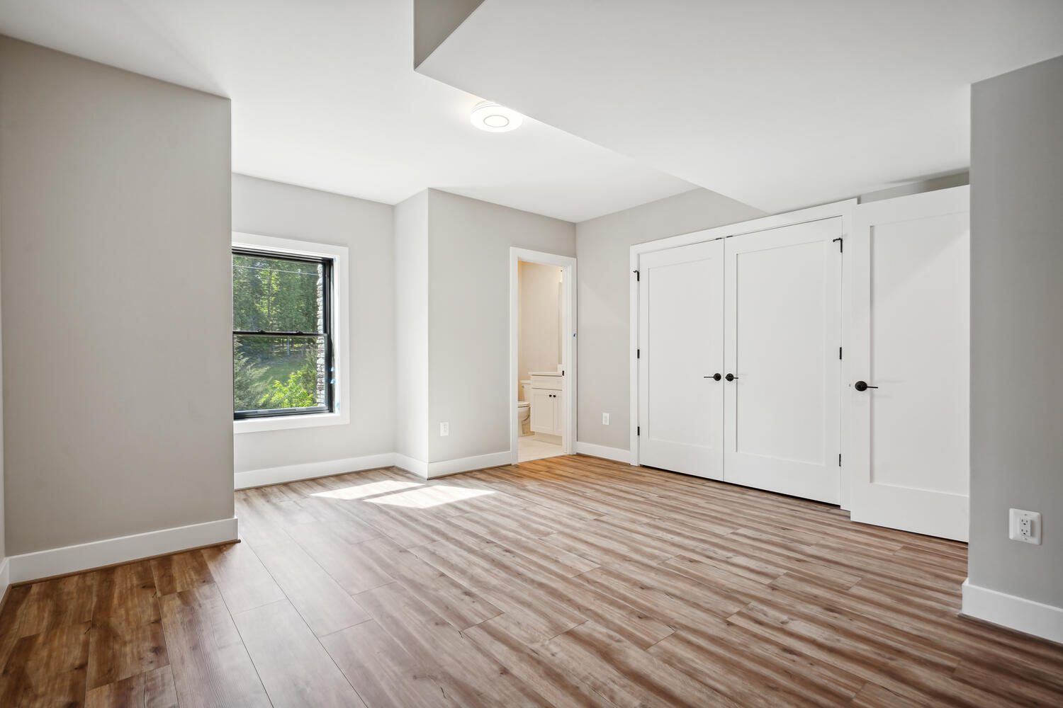 Empty room with light wood flooring, white walls, a window on the left, two white double doors on the right, and a doorway leading to a small bathroom.