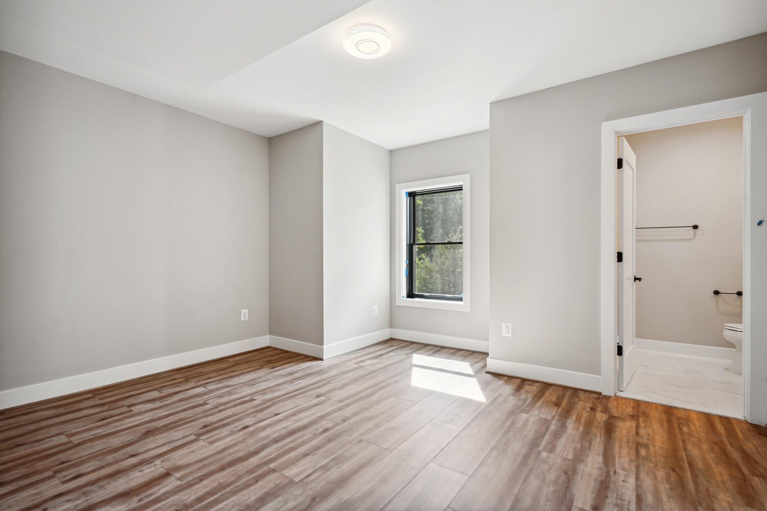 An empty room with wooden floors, a single window, and gray walls. A door to the right leads to a small bathroom with white tiles and a toilet.