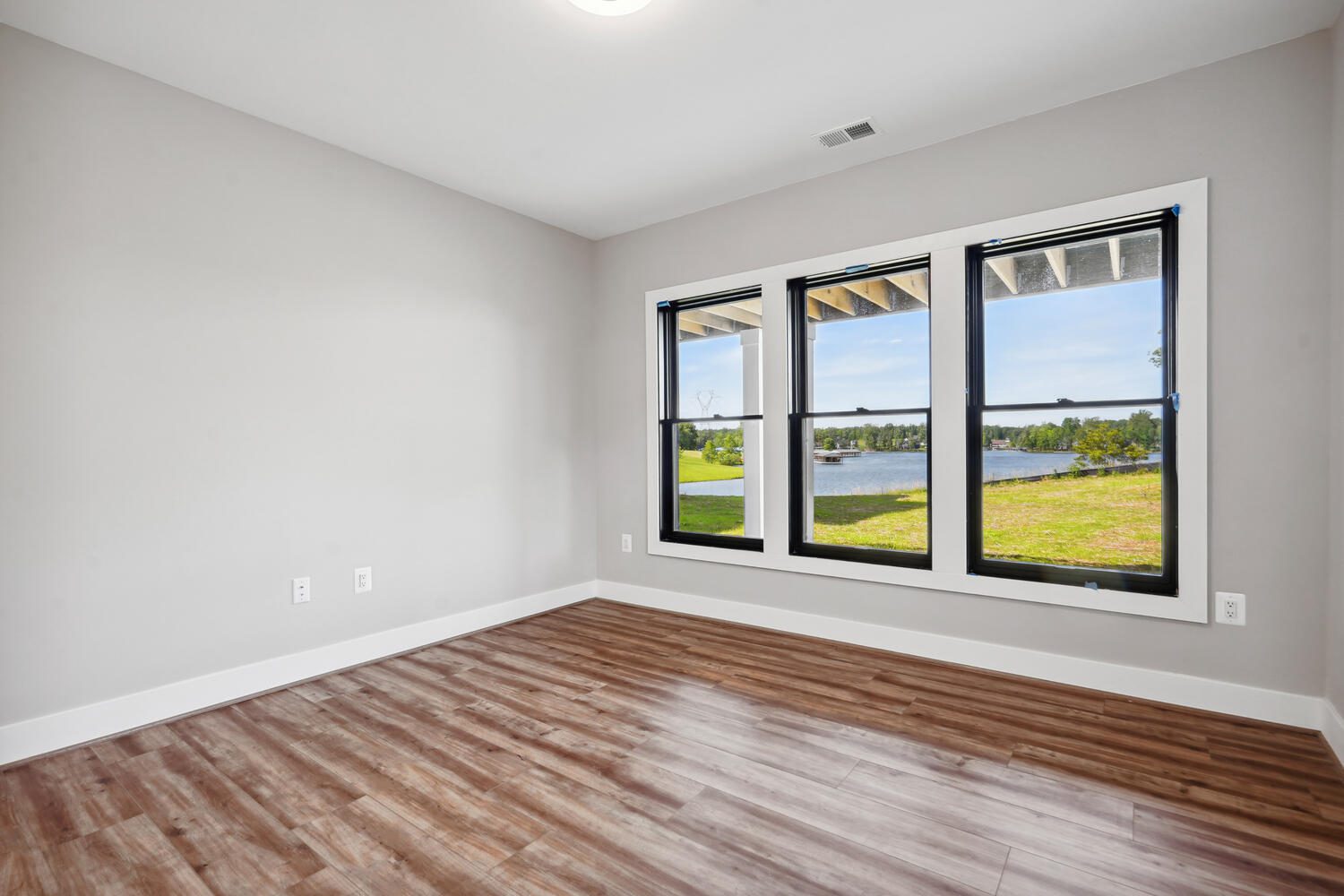 An empty room with wood flooring, beige walls, and three large windows overlooking a lake and grassy area.