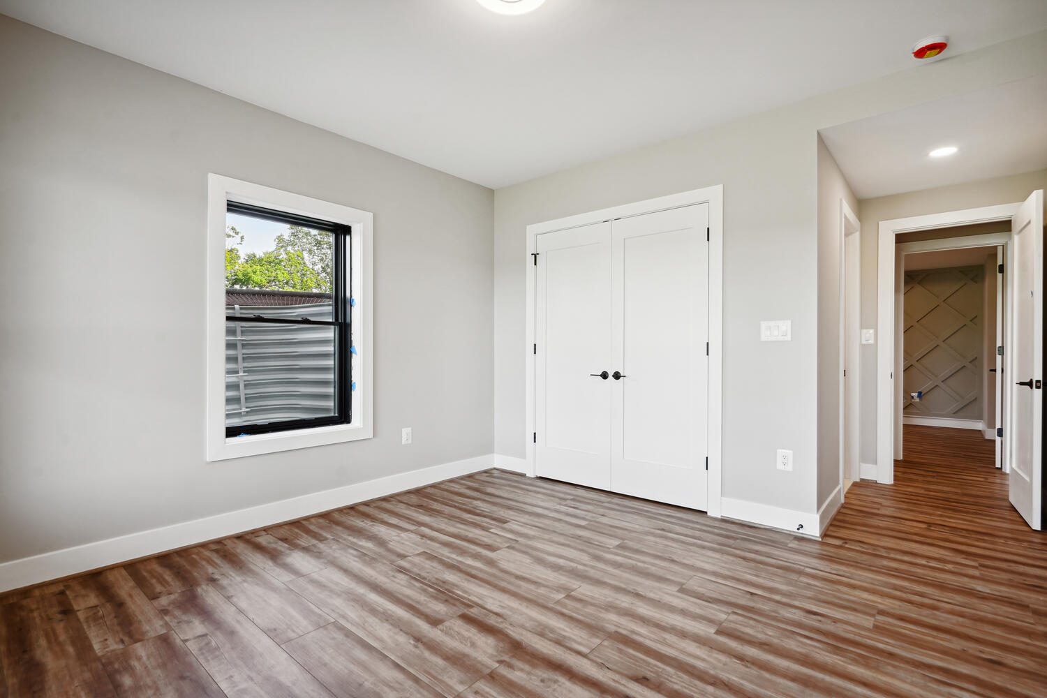 Empty room with wood flooring, a large window on the left, and white double doors on the right. Hallway visible through an open door. Walls are light gray.