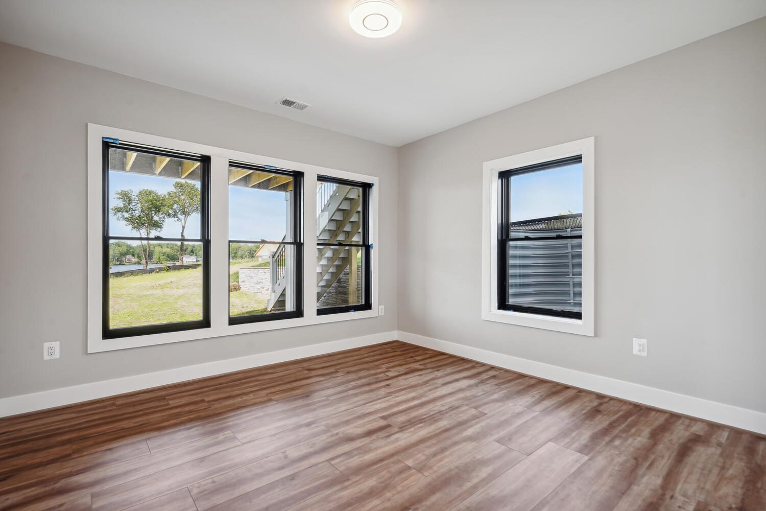 Empty room with light wood flooring, gray walls, and a ceiling light. Large windows on the left and a smaller window on the right offer views of a grassy yard and trees outside.