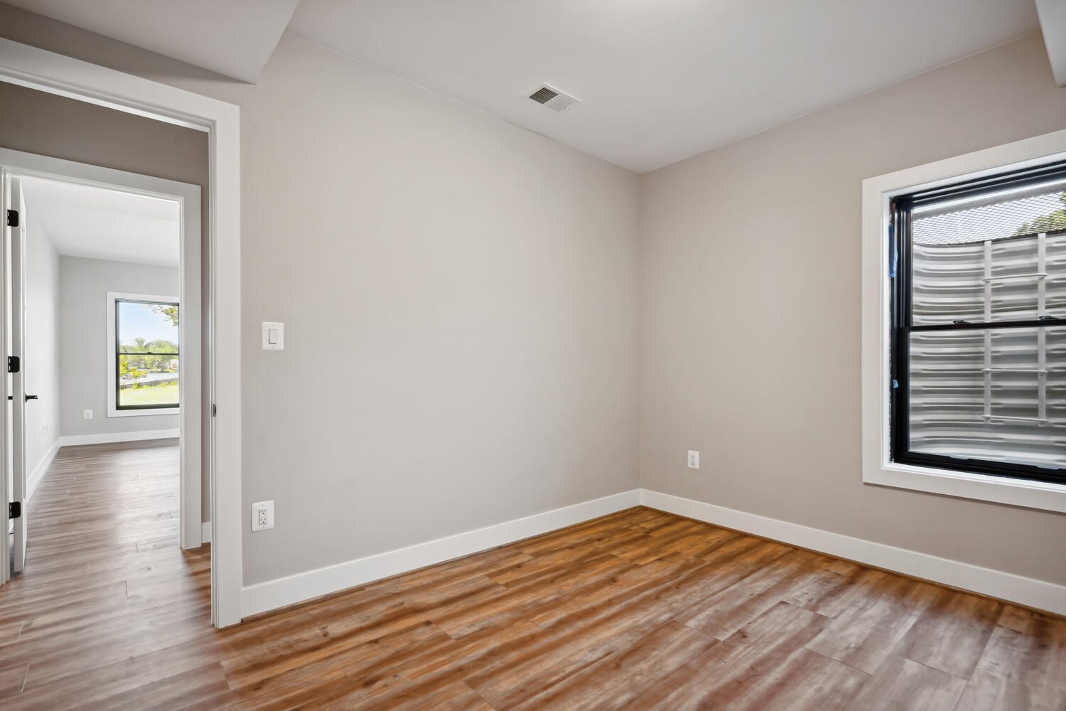 Empty room with light wood flooring, beige walls, a window with a cover, and an open door leading to another room with a window.