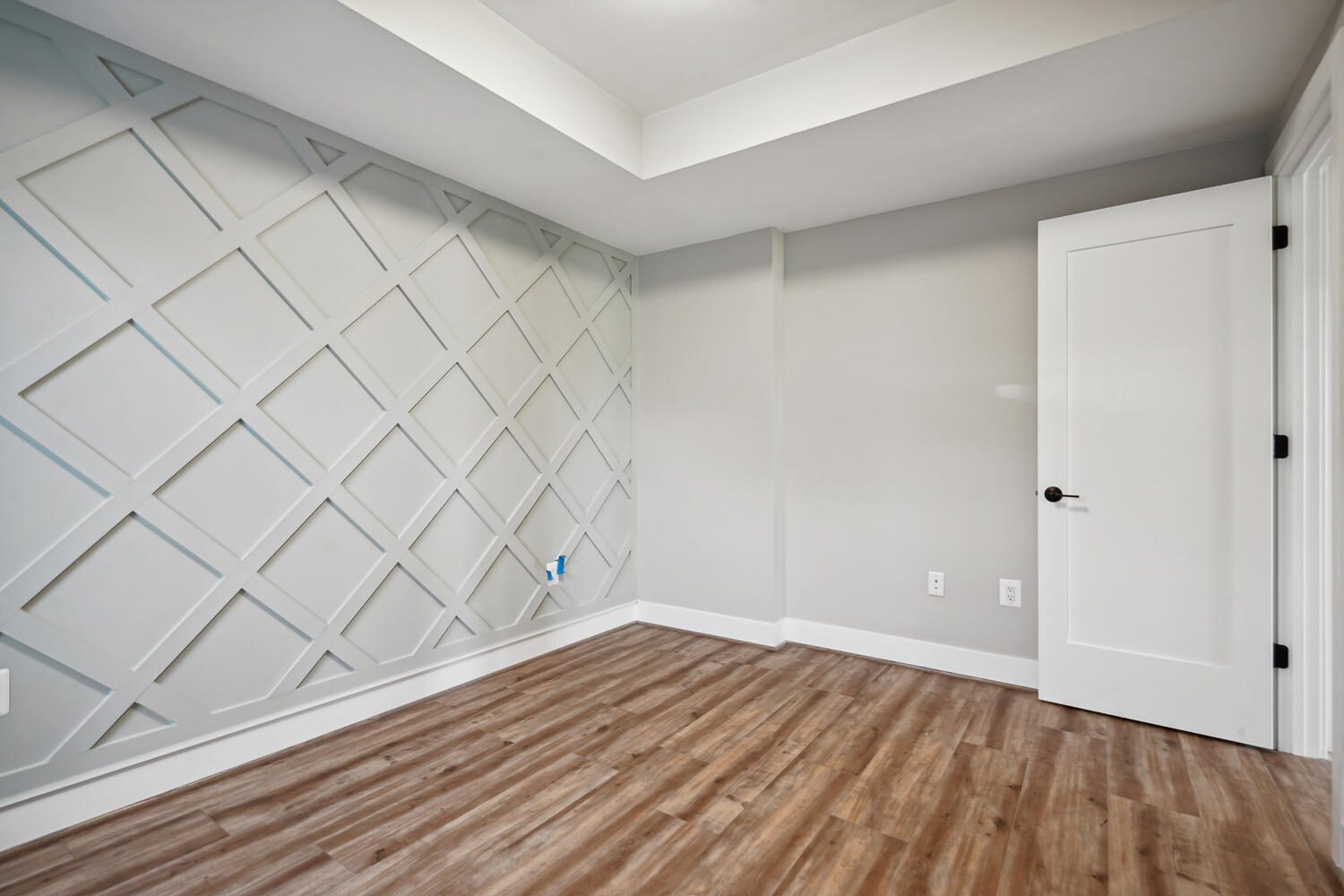 Empty room with light gray walls, a geometric accent wall, wood flooring, and a white door.