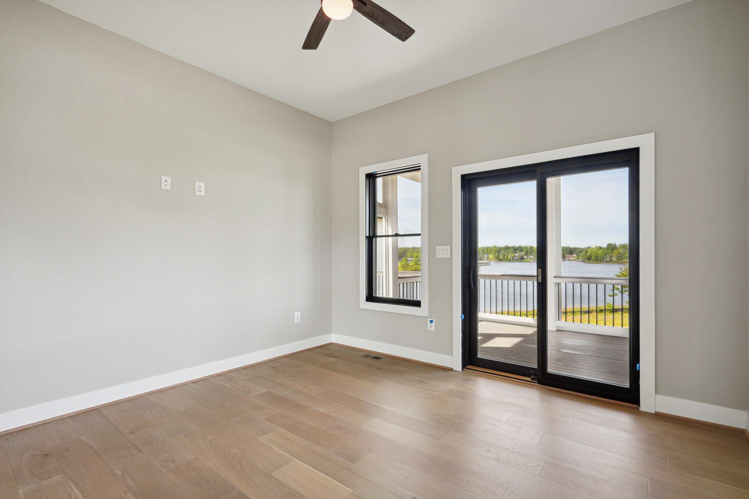 Empty room with light wood floors, beige walls, a ceiling fan, and large glass doors leading to a balcony overlooking a lake or river.