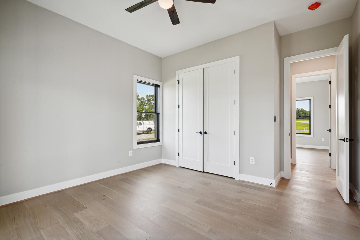 Empty room with light wood flooring, white walls, a ceiling fan, two doors, and a window showing an outdoor view.