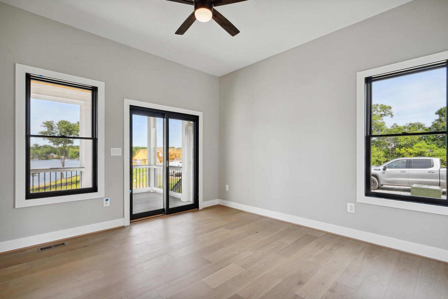 Empty room with light wood flooring, gray walls, ceiling fan, and large windows. Glass doors open to a small balcony with a view of a lake and a parked truck outside.