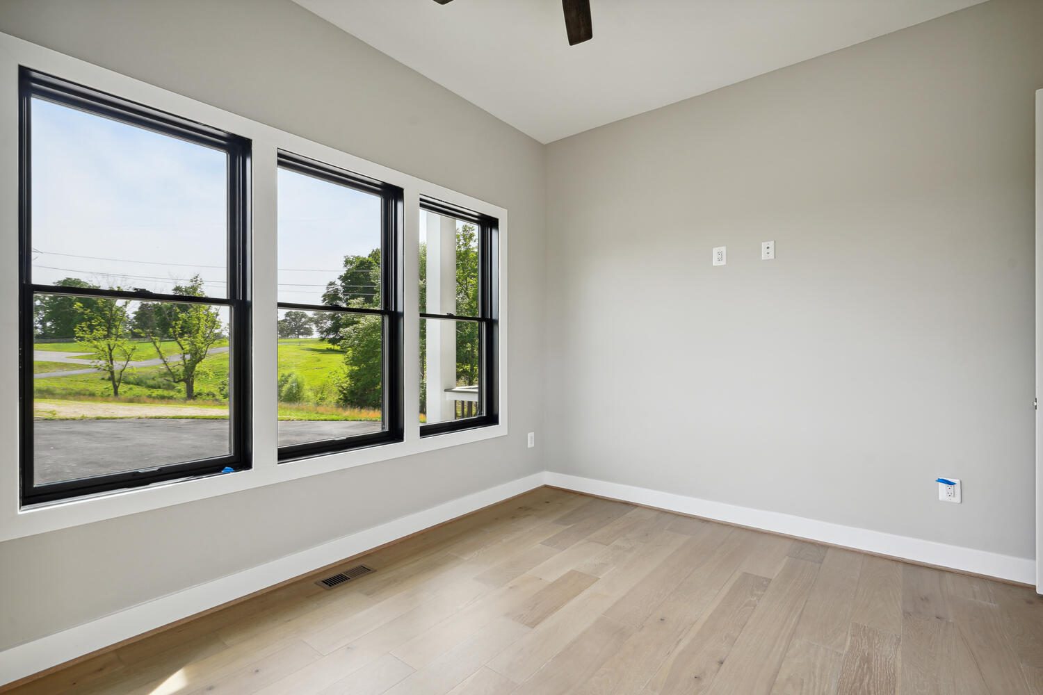 Empty room with light wood flooring, gray walls, and a ceiling fan. Three large windows offer a view of greenery and a paved road outside. Electrical outlets visible on walls.