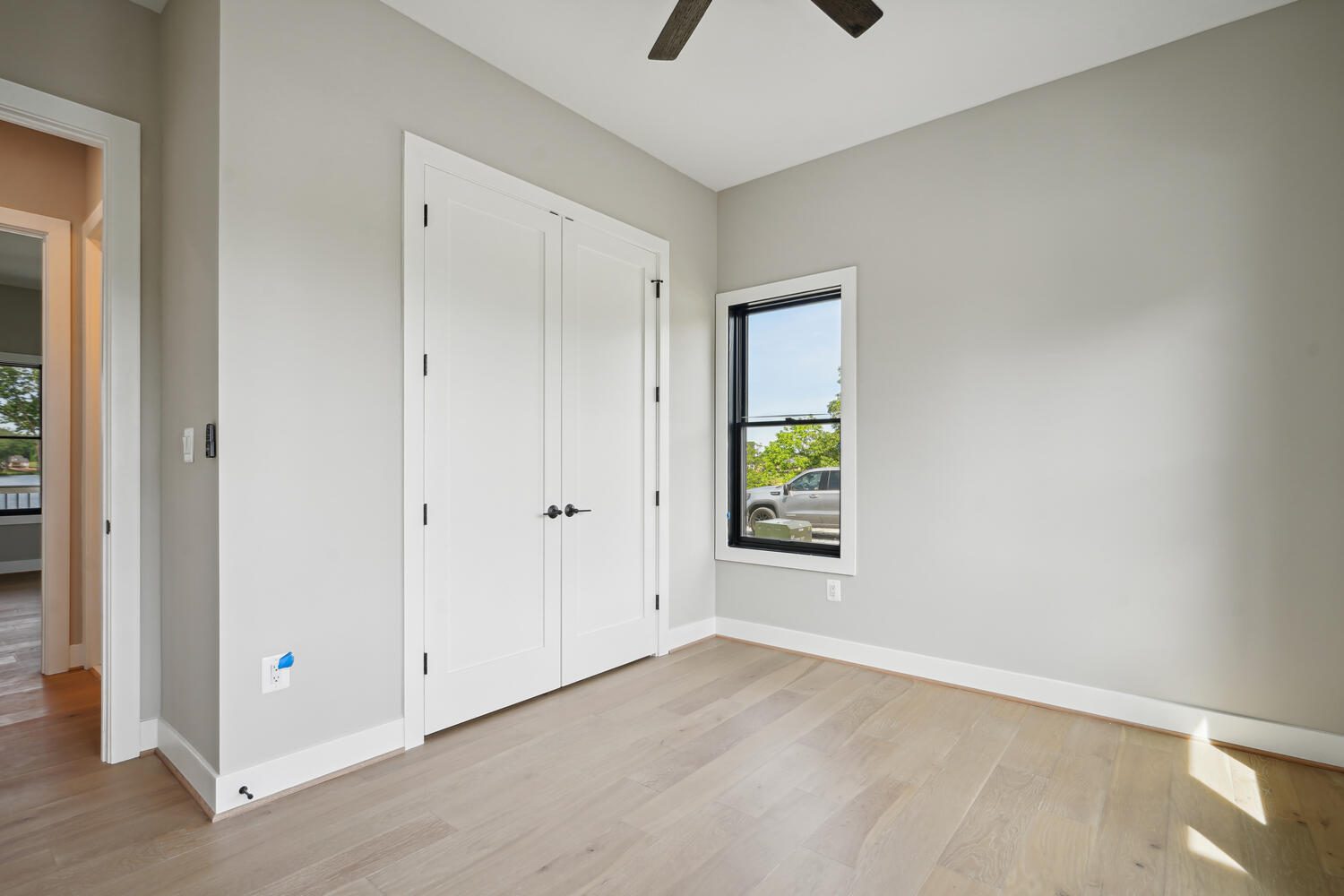 Minimalist empty room with light wood flooring, beige walls, a ceiling fan, a double door, and a window with a view of trees and a parked car outside.