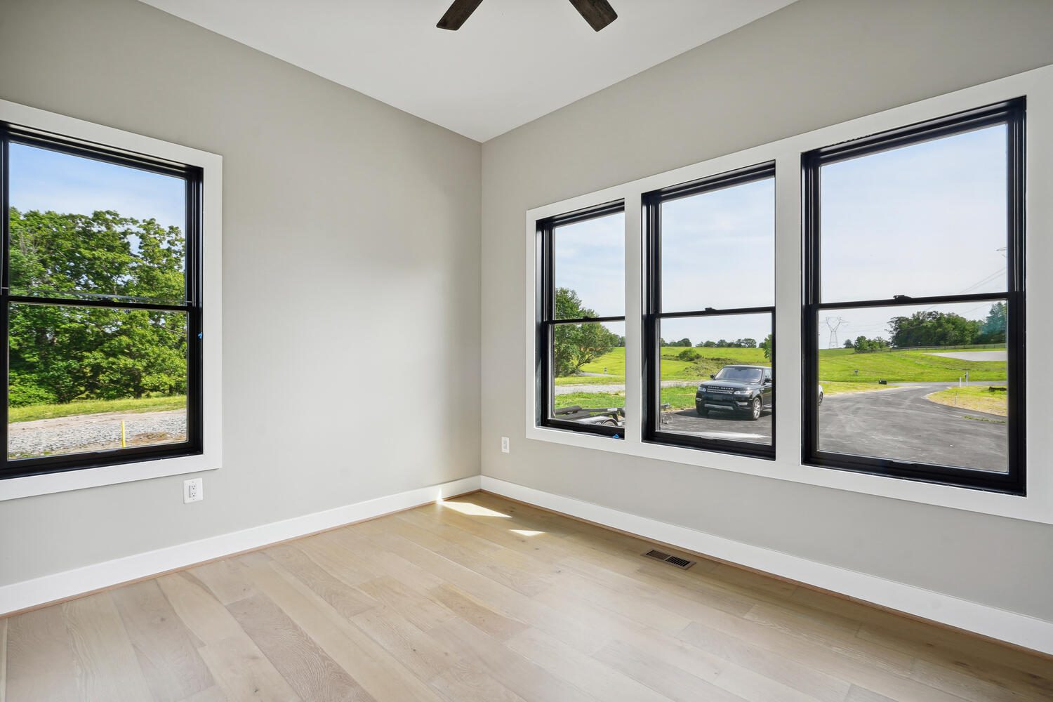 Empty room with light wood flooring and gray walls, featuring three large windows with black frames. Outside, a car is parked on a paved road with grass and trees in the background.