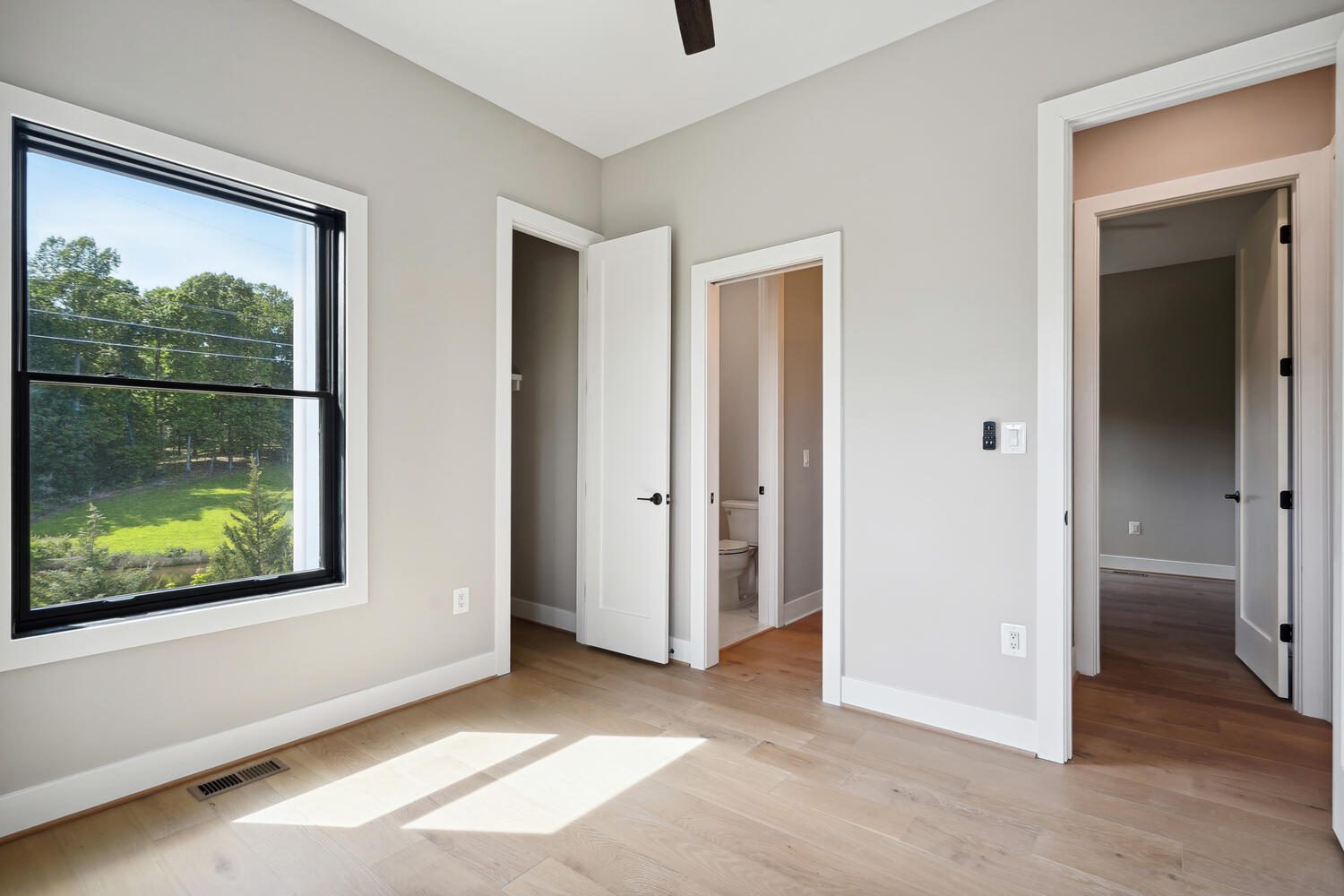 Empty room with light wood flooring, large window, two open white doors, and a small closet. The window shows trees outside, and a ceiling fan is partially visible.