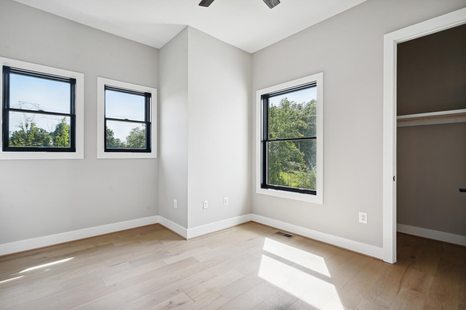 Empty room with light wood flooring, gray walls, and three black-framed windows. A small open closet is on the right, and the room is sunlit.