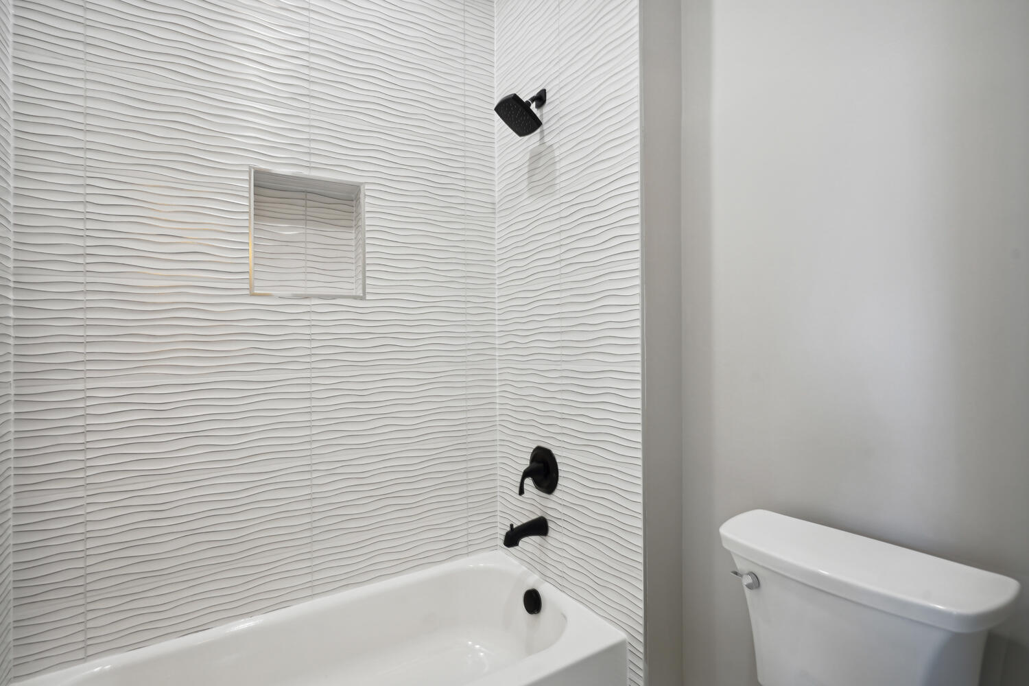 A bathroom with a white bathtub, wavy textured white tile wall, built-in shelf, black fixtures, and a white toilet.