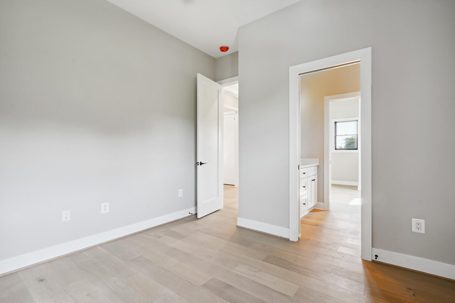 An empty room with light wood flooring, gray walls, an open white door leading to a bathroom, and natural light coming through a window.