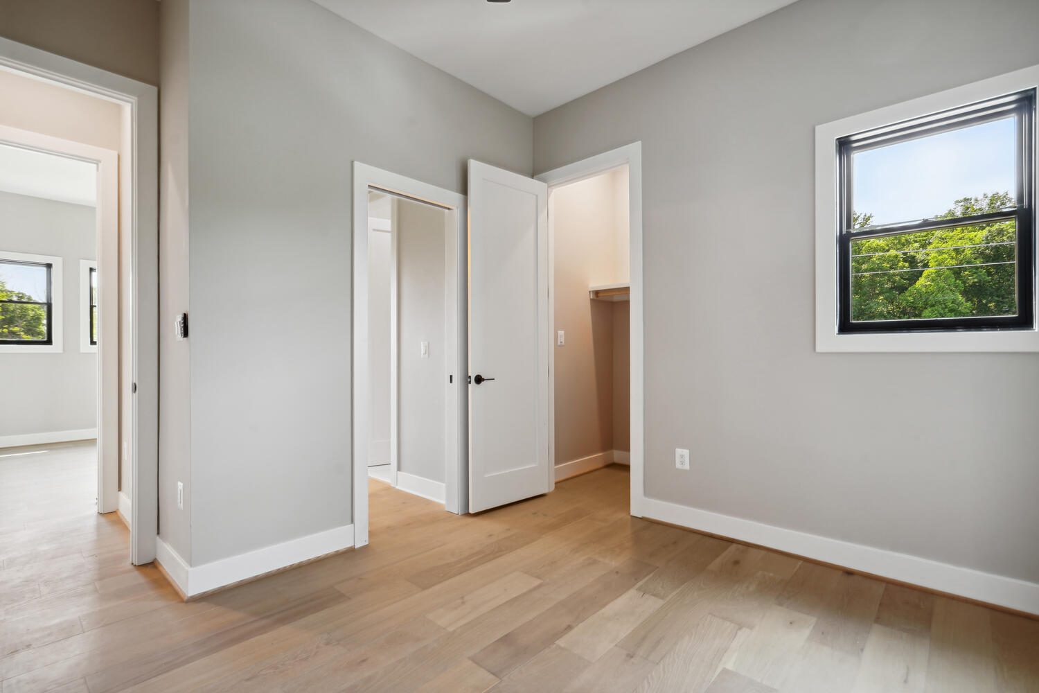 Empty room with light wood floors, beige walls, a window showing greenery, a white door to a closet, and another open door leading to an adjoining room.