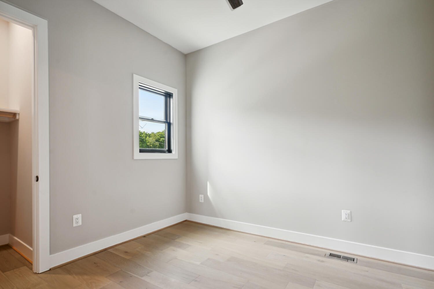 Empty room with light wood flooring, a small window with a view of greenery, and an open closet door on the left. Walls are painted light gray with a white ceiling.