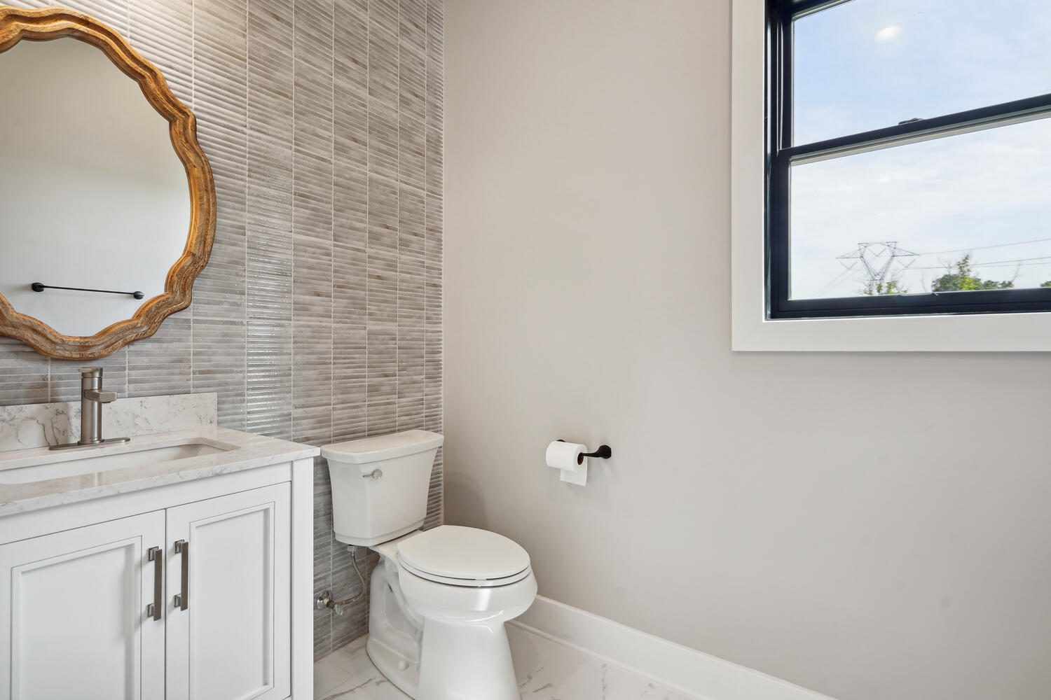 Bathroom with a white toilet, marble countertop, round mirror, and gray tiled wall. A window on the right provides natural light.