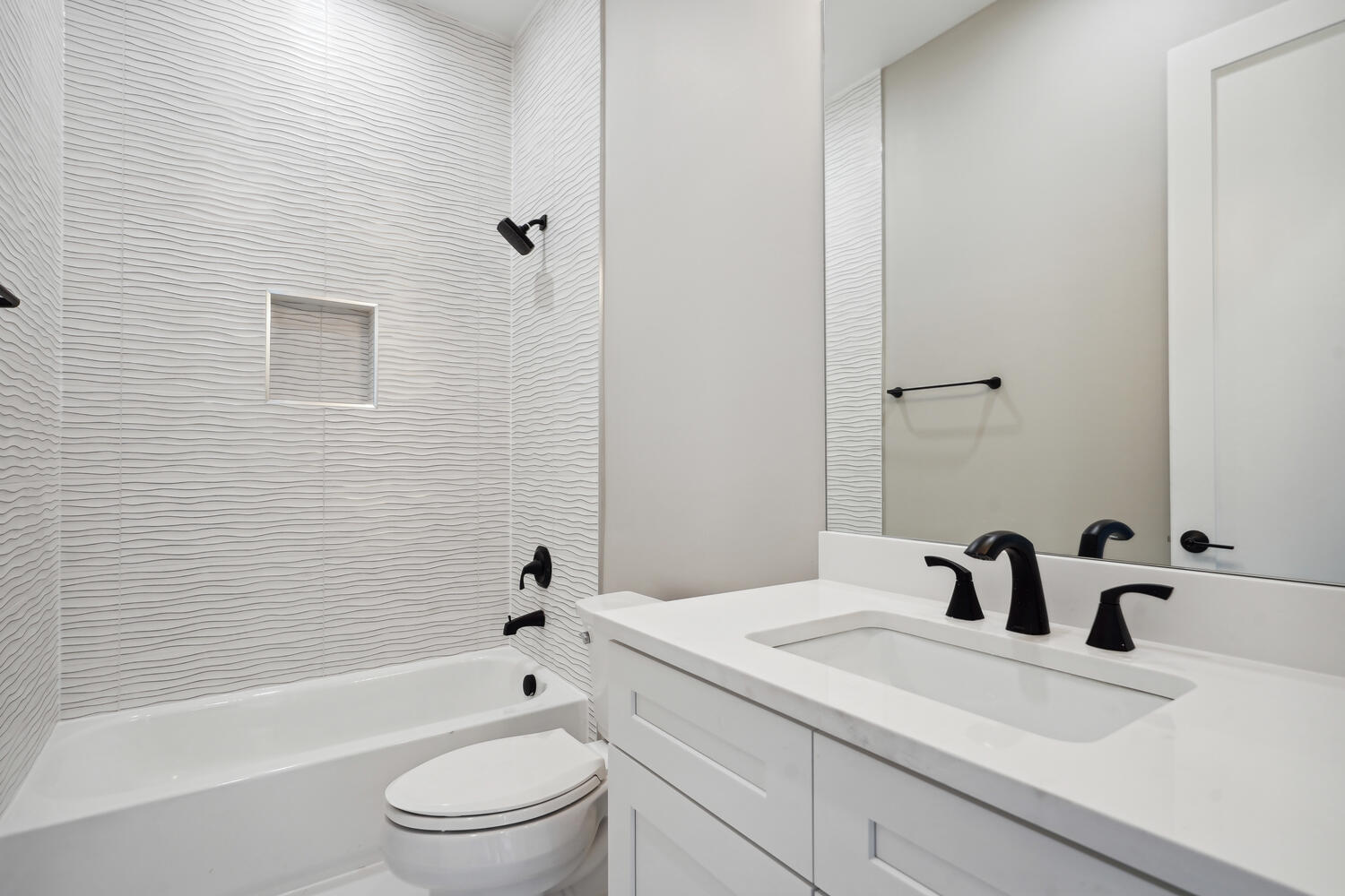 A modern bathroom featuring a white bathtub, a black showerhead, a toilet, and a white vanity with a black faucet. The walls have a textured design, and a large mirror is above the sink.