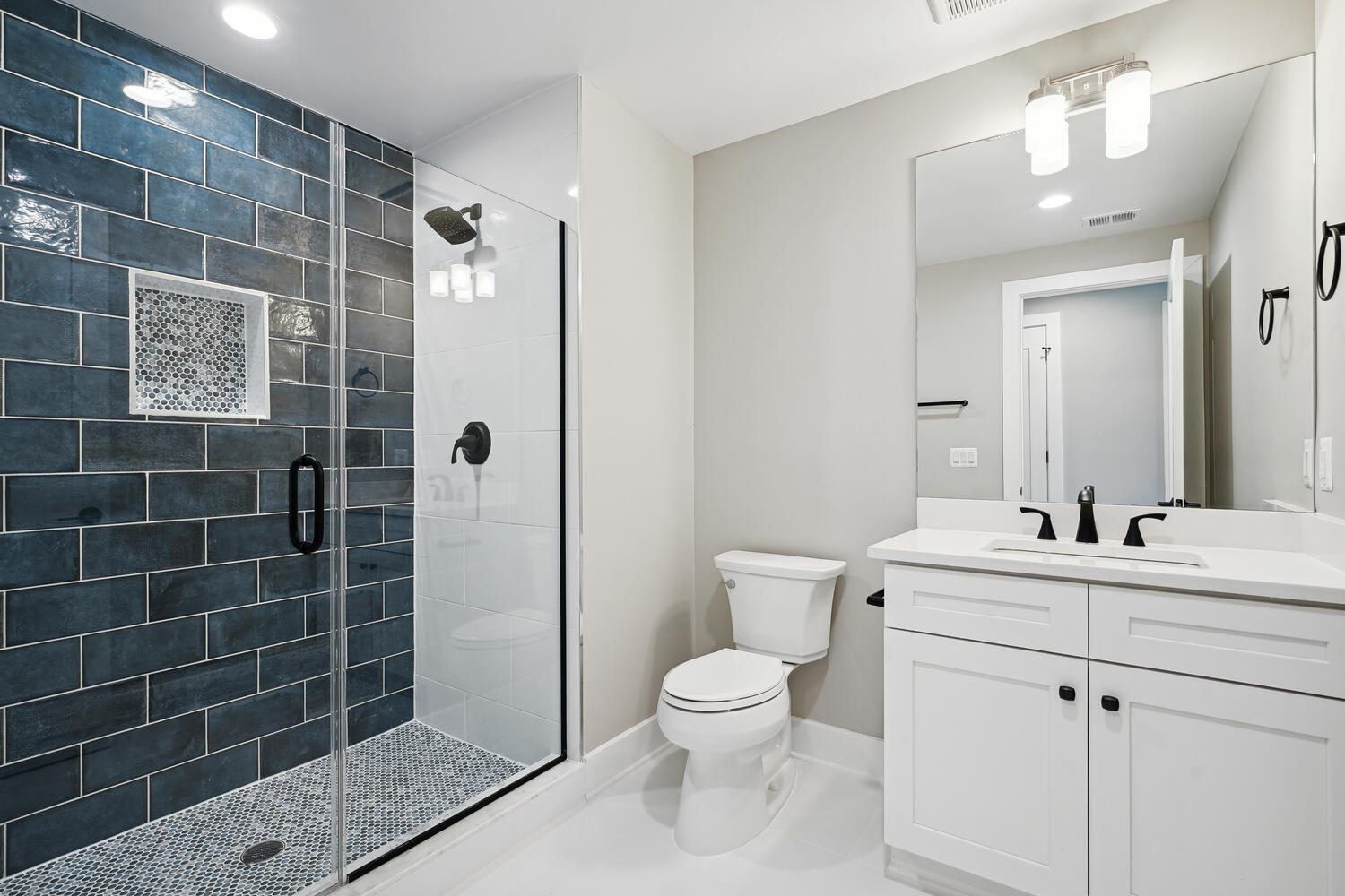 Modern bathroom with a glass-enclosed shower featuring dark blue tiles, a white vanity with a mirror above, and a toilet. Bright lighting from ceiling fixtures illuminates the space.