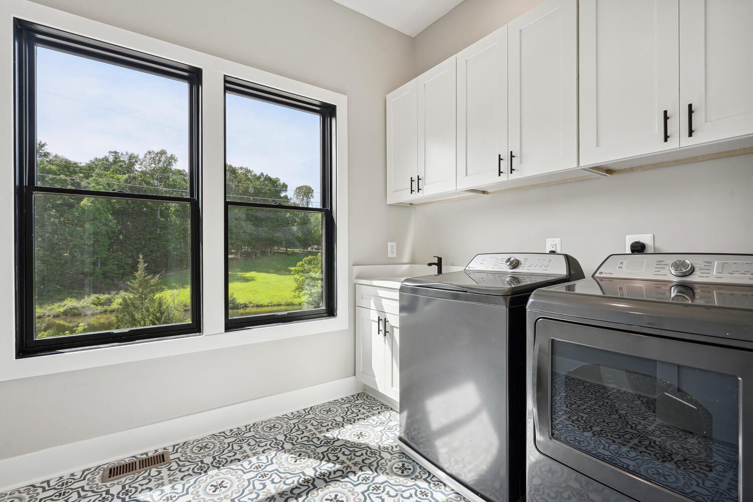 Laundry room with a black washer and dryer, white cabinets, a sink, and a patterned tile floor. A large window offers a view of greenery outside.