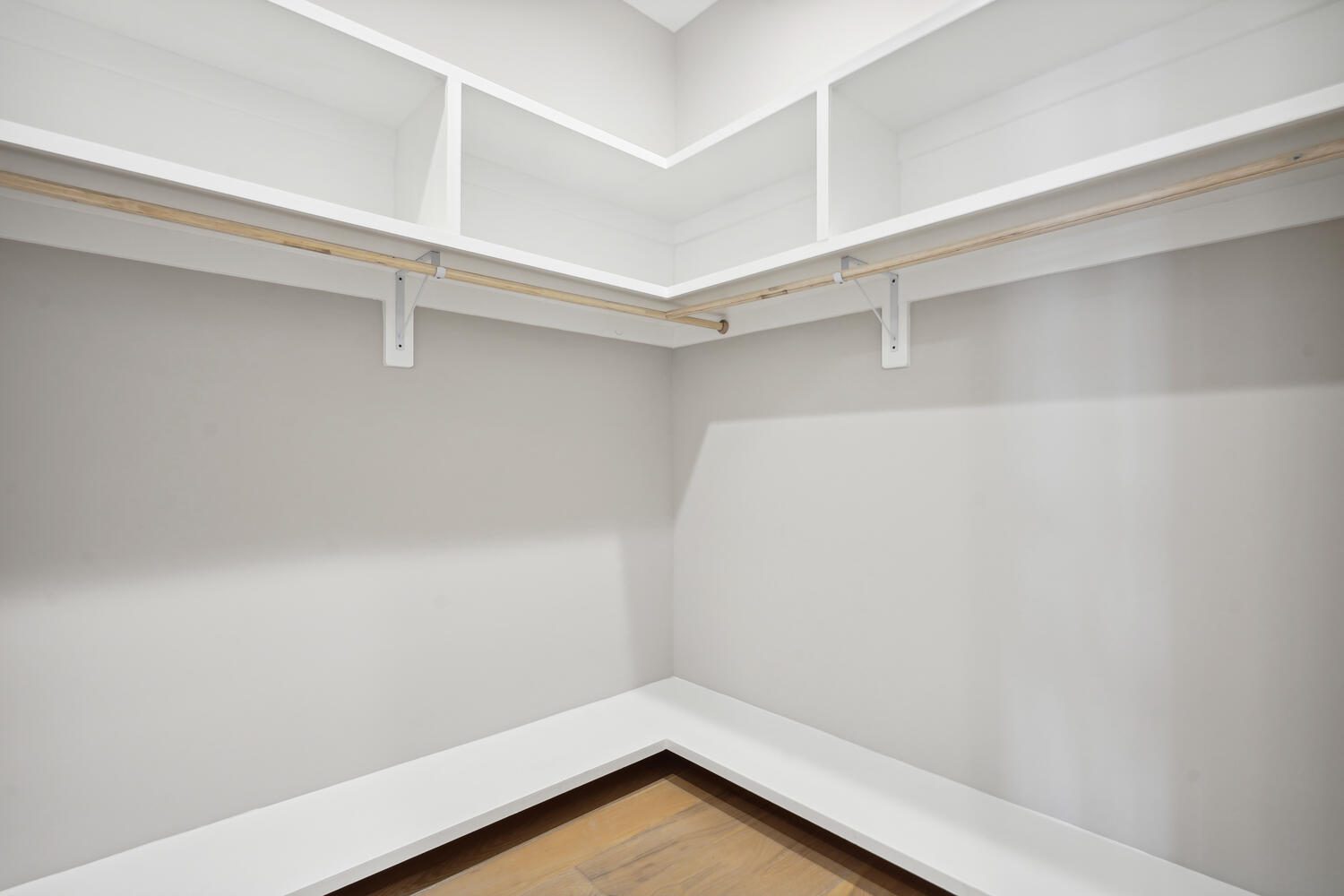 Empty white walk-in closet with wooden shelves and rods against light gray walls, and wooden floor.