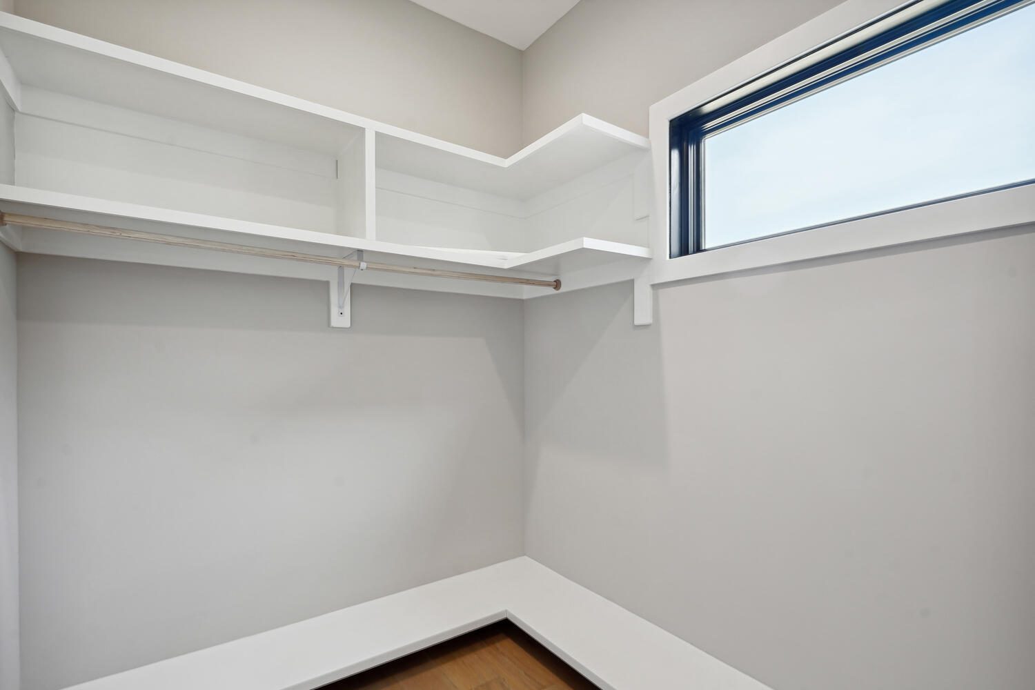 Empty corner closet with white shelves, a clothing rod, and a low bench. Light gray walls and a rectangular window provide natural light.