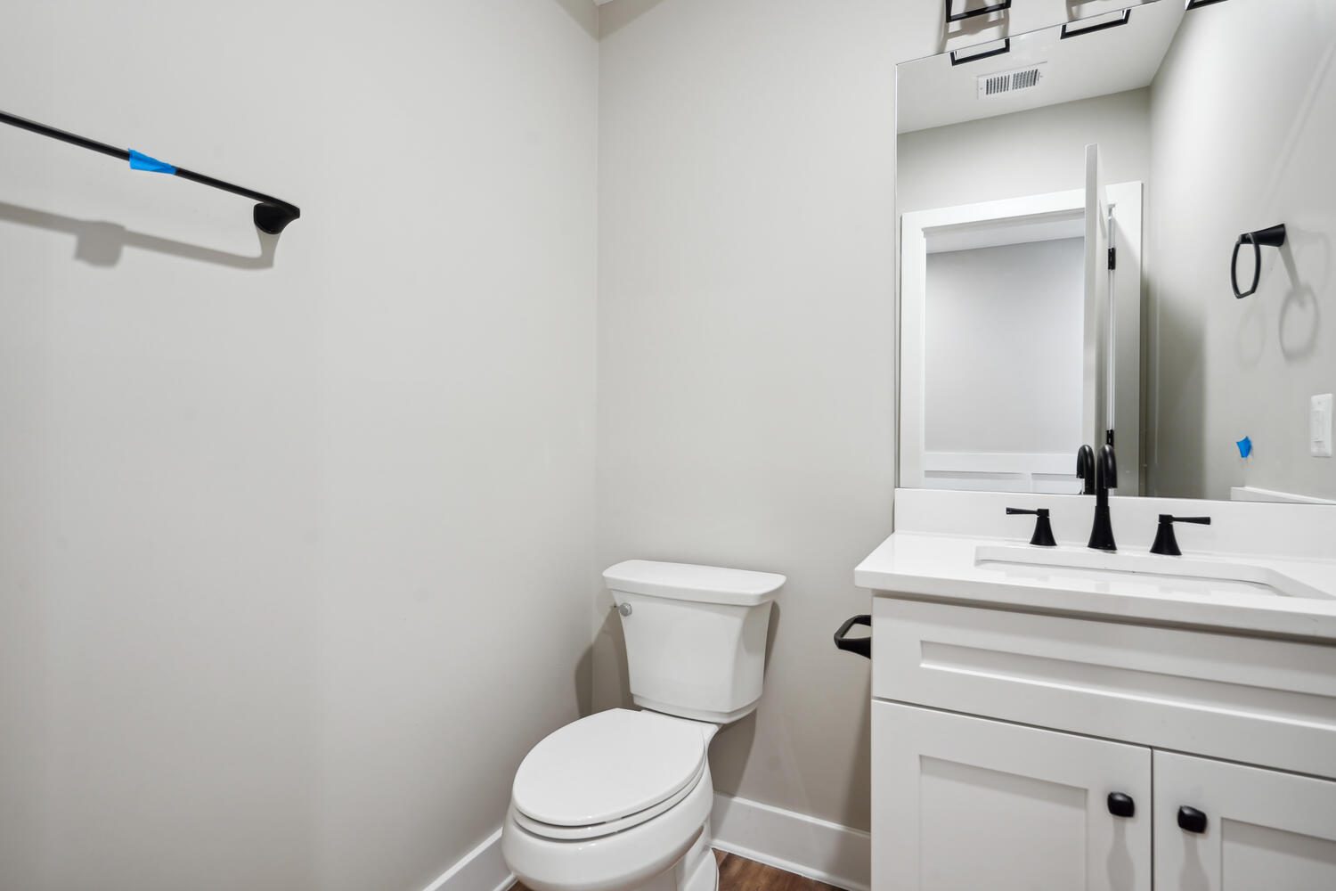 Small bathroom with a white toilet and vanity, a black faucet and hardware, a mirror above the sink, and a towel rod on the wall. Walls are light gray, and the floor is wooden.