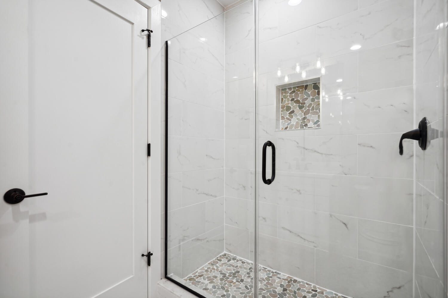 A white-tiled shower with a glass door and black handles, featuring a built-in niche and a pebble stone floor inlay.