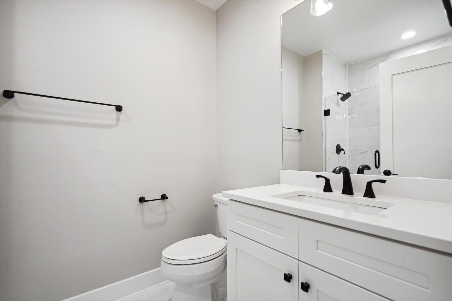 Minimalist bathroom with white vanity, black fixtures, a toilet, and a shower. Neutral wall and floor colors complement the simple design.