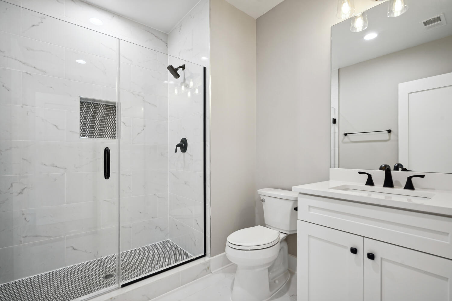 Modern bathroom with a glass-enclosed shower, white vanity with twin faucets, white countertop, and a toilet. Walls and floor are tiled in light tones.