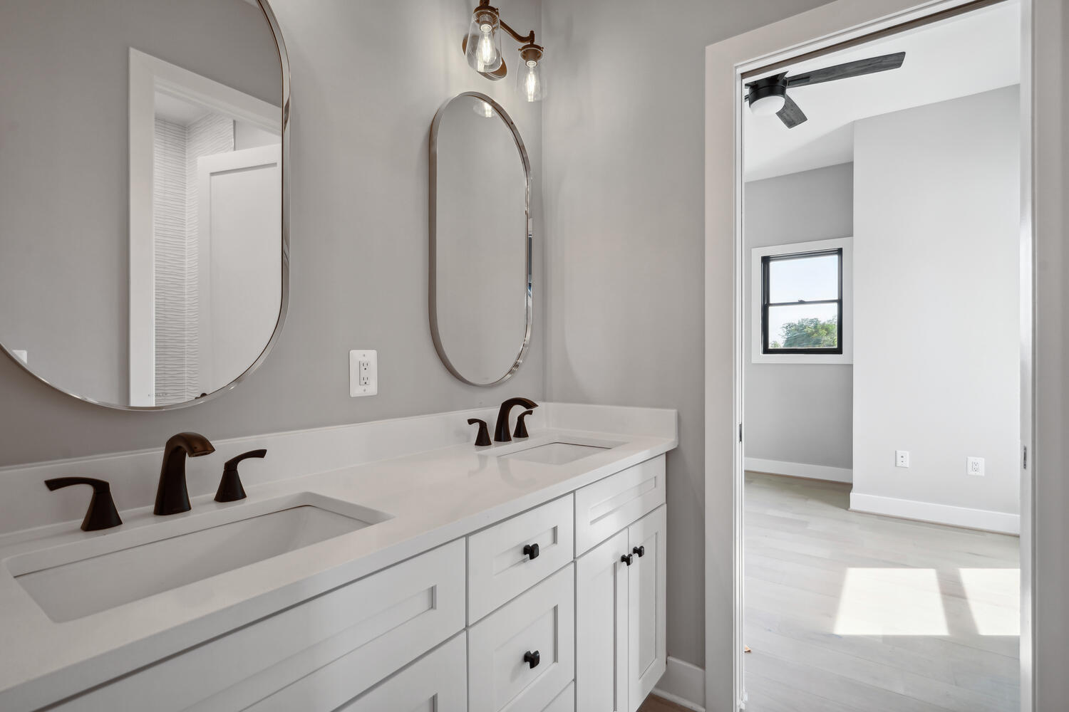 A modern bathroom with two oval mirrors above a double sink vanity, black faucets, white cabinetry, and a view into an adjacent room with a window and ceiling fan.