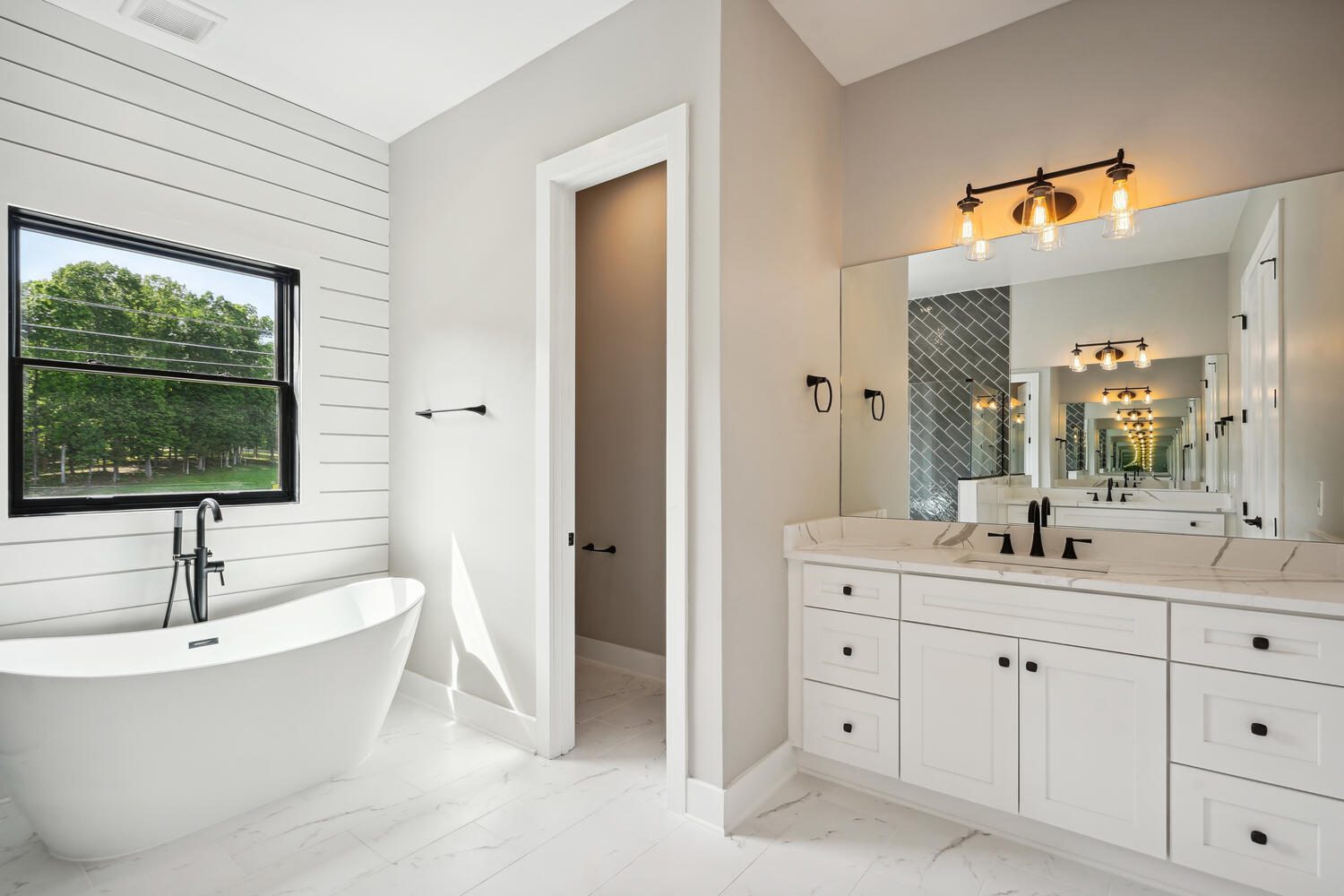Modern bathroom with a freestanding bathtub, black faucet, large window, vanity with double sink, mirror, and light fixture. Walls are painted light gray with some shiplap siding.