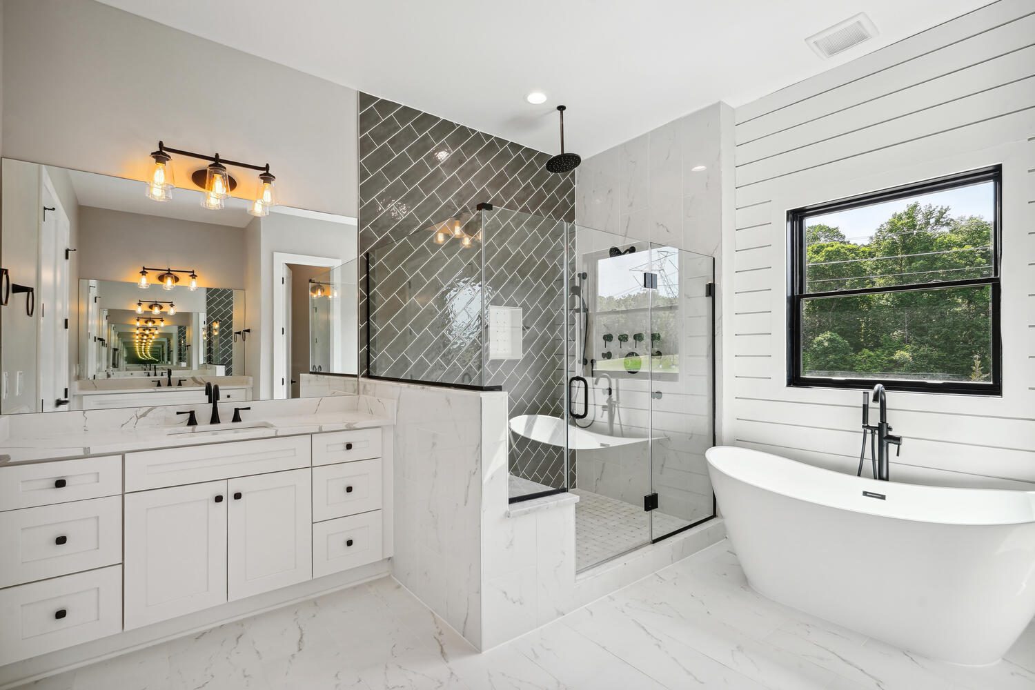 Modern bathroom with white cabinets, dual sinks, glass shower, freestanding tub, black fixtures, and a window overlooking greenery. Gray herringbone and white shiplap walls add texture.