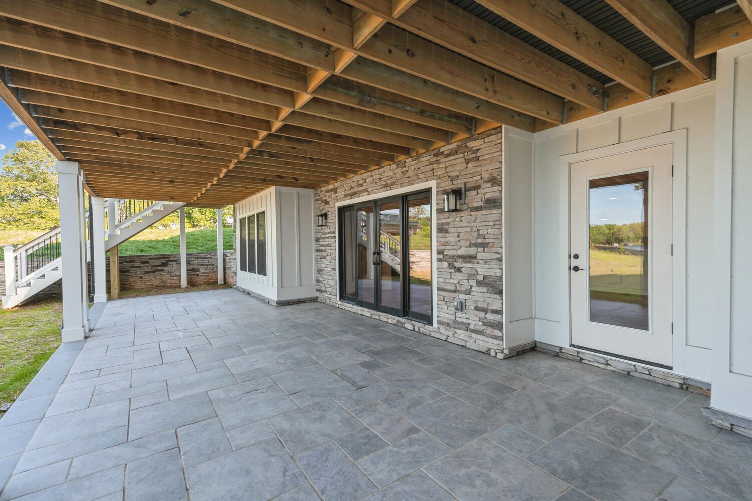 Covered patio with stone flooring, glass doors, and exposed wooden beams. Stairs lead to the yard, and the space is bordered by stone and white siding walls.