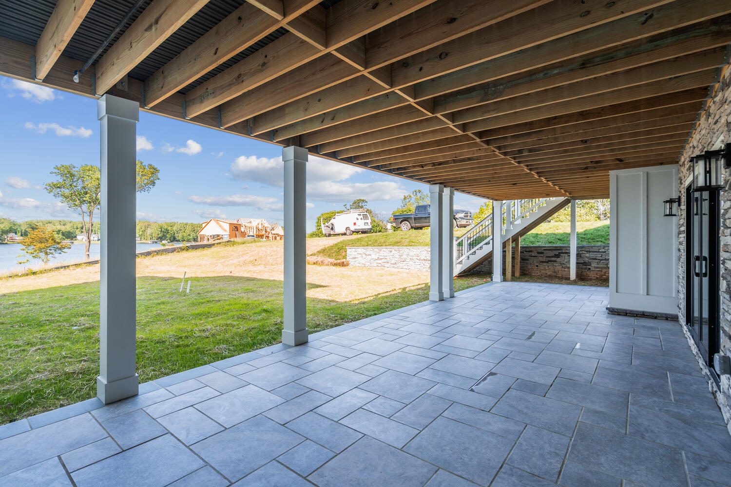 Covered patio with stone tiles, supported by wooden beams, overlooks grassy area and lake. Stairs lead to the upper level.