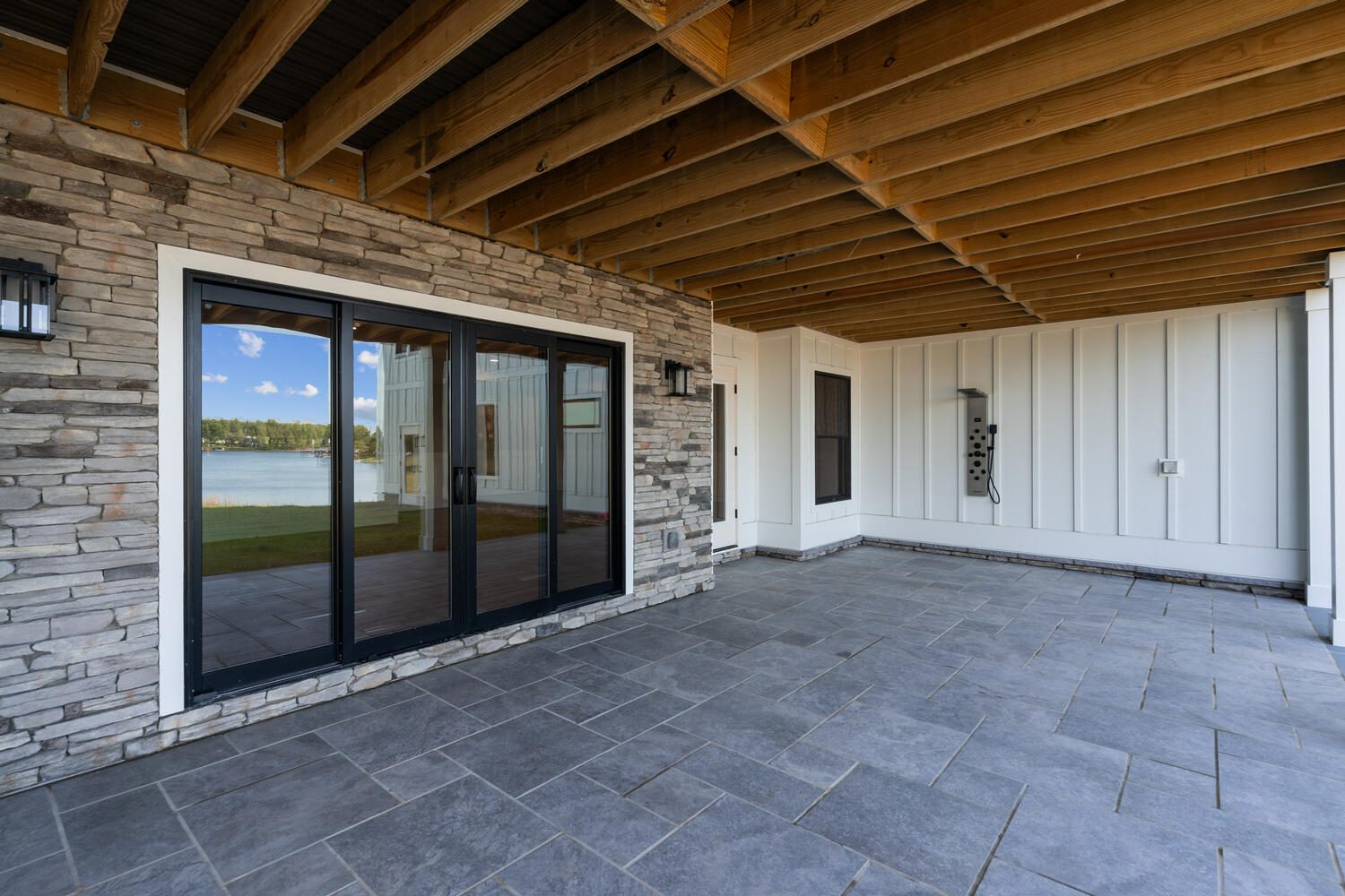 Covered patio with stone walls and large glass doors, overlooking a lake. The ceiling features exposed wooden beams, and the floor is tiled with gray stones.