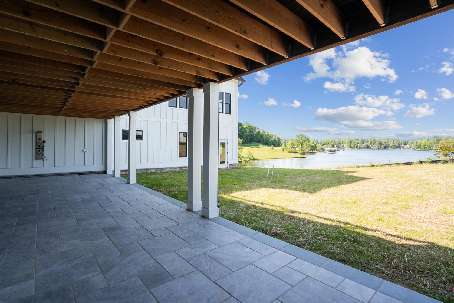 Covered patio with gray tiles beneath a wooden ceiling, overlooking a grassy area and a lake with trees and blue sky in the background.