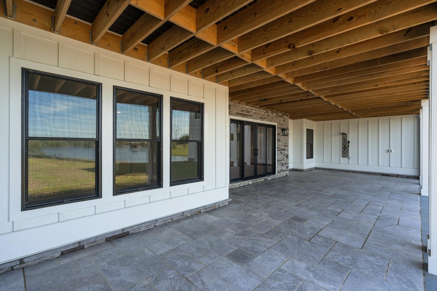 Covered patio with a tiled floor, wooden ceiling beams, and large windows. Stone accent wall and sliding glass door on the right. Scenic view visible through the windows.