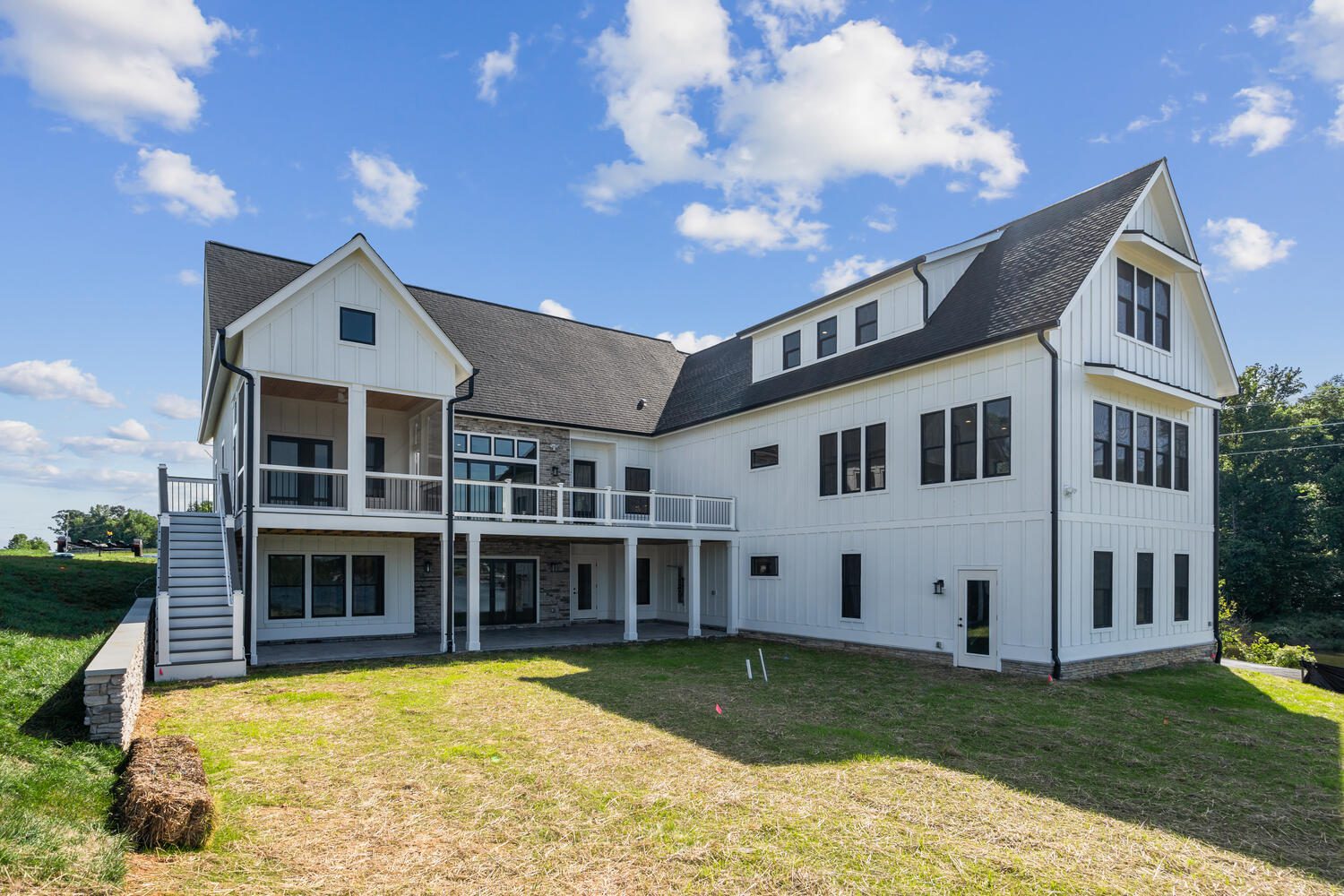 Large modern white house with dark roof, multiple windows, and a covered porch. Grass yard with some bare patches under a blue sky with few clouds.