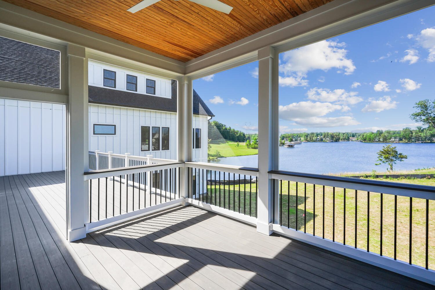 Covered porch with wooden ceiling and railing overlooks a lake and greenery, attached to a white house.