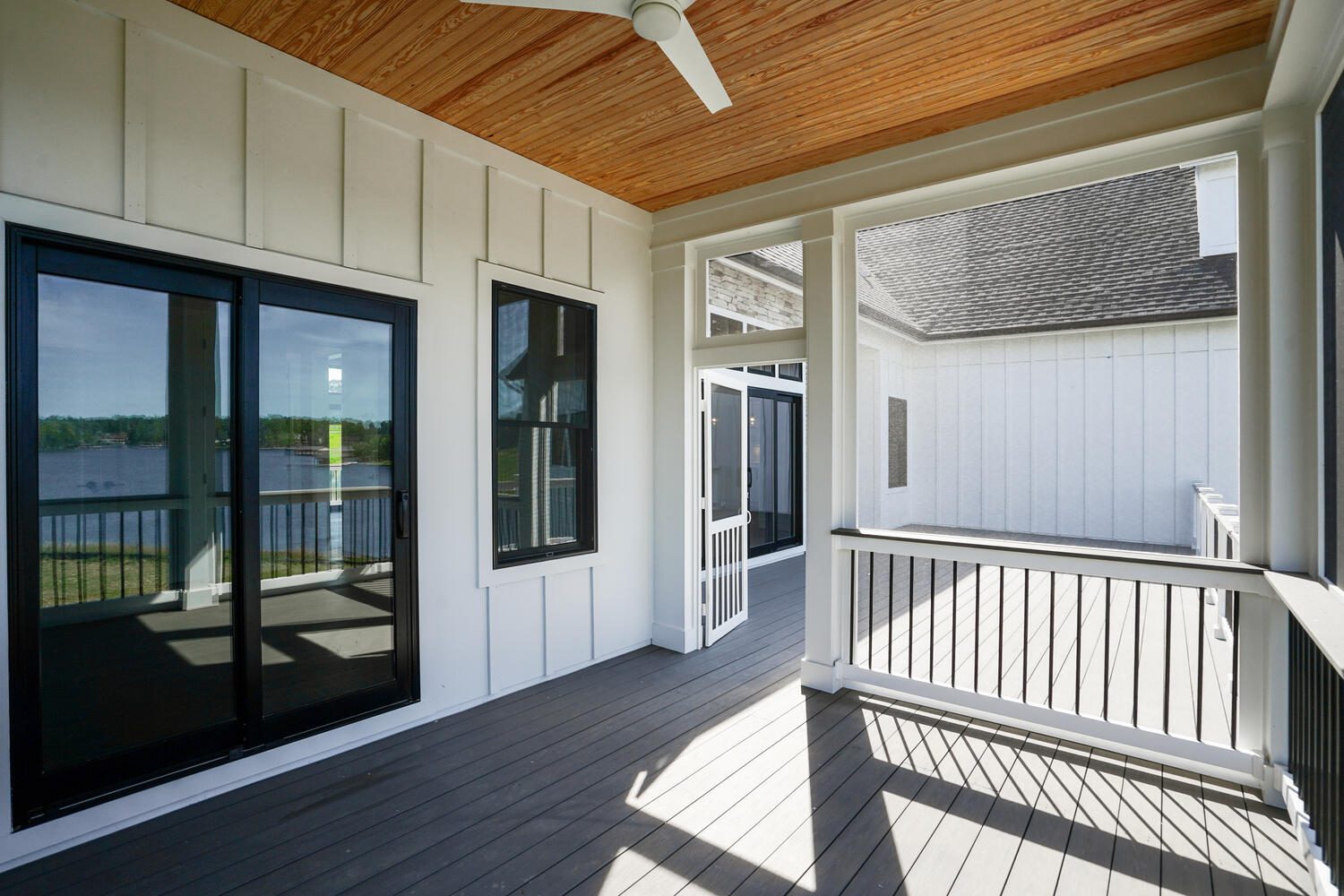 Screened porch with wood ceiling, dark-framed windows, and gray flooring. Overlooks a lake and connects to an open deck with white railing. Ceiling fan is visible above.
