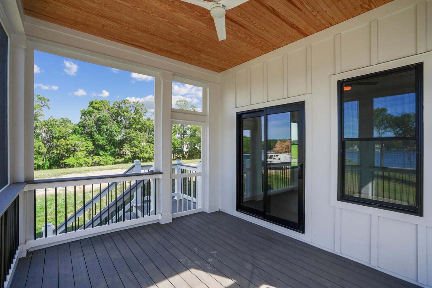 A wooden porch with a ceiling fan, featuring a railing overlooking a grassy area. Sliding glass doors and windows are seen on the right.