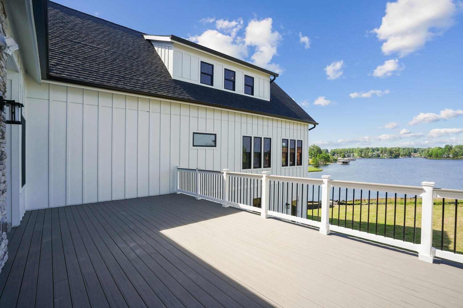 A house with white siding and a dark roof has a spacious deck overlooking a lake. The sky is clear with a few clouds.