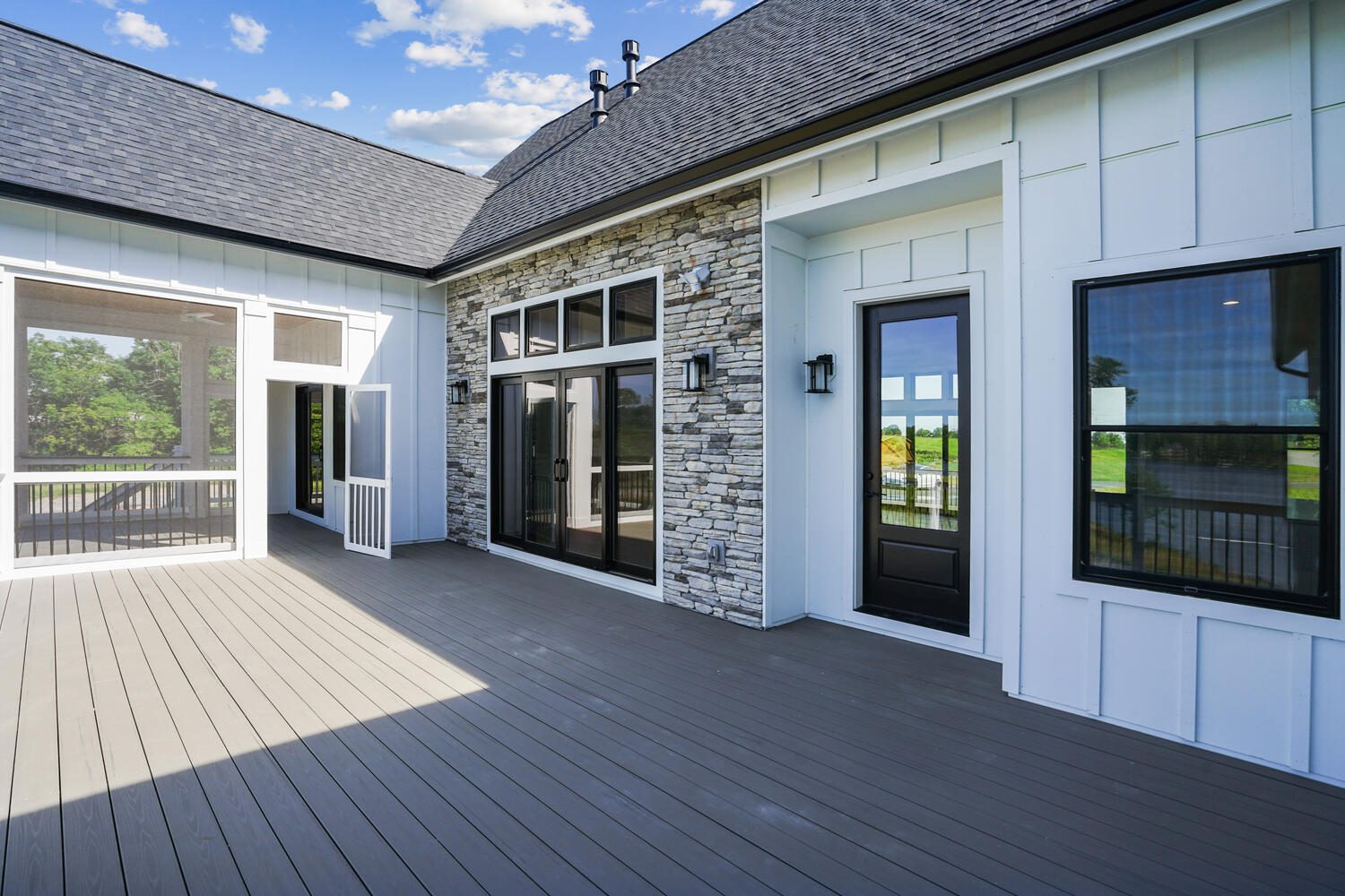 Spacious patio with wooden flooring, stone accent wall, black-framed windows, and doors. Blue sky with scattered clouds is visible in the background.
