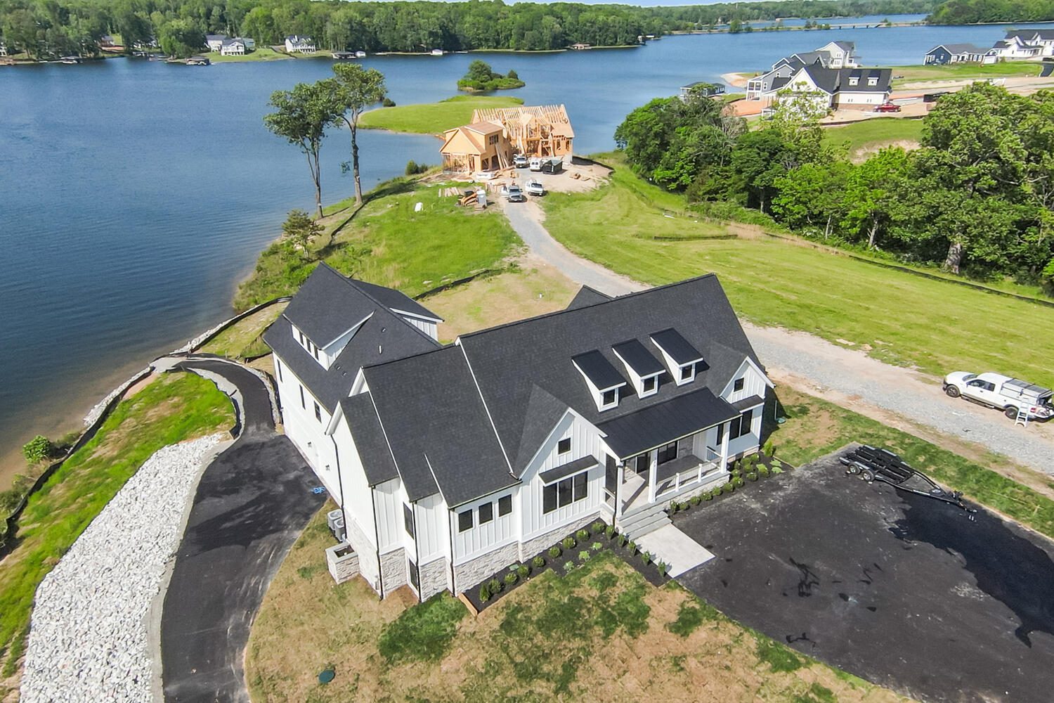 Aerial view of a modern white house with a black roof, situated near a lake. There's a paved driveway, surrounding greenery, and another house under construction nearby.