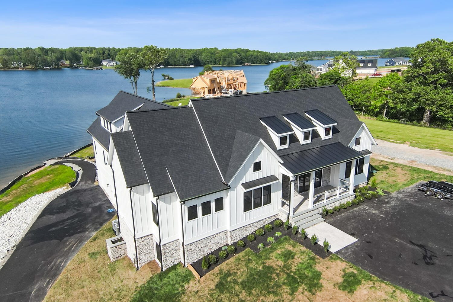 Large white lakeside house with black roof, multiple dormer windows, and a manicured lawn. Surrounded by water and greenery under a clear blue sky. Several houses nearby.