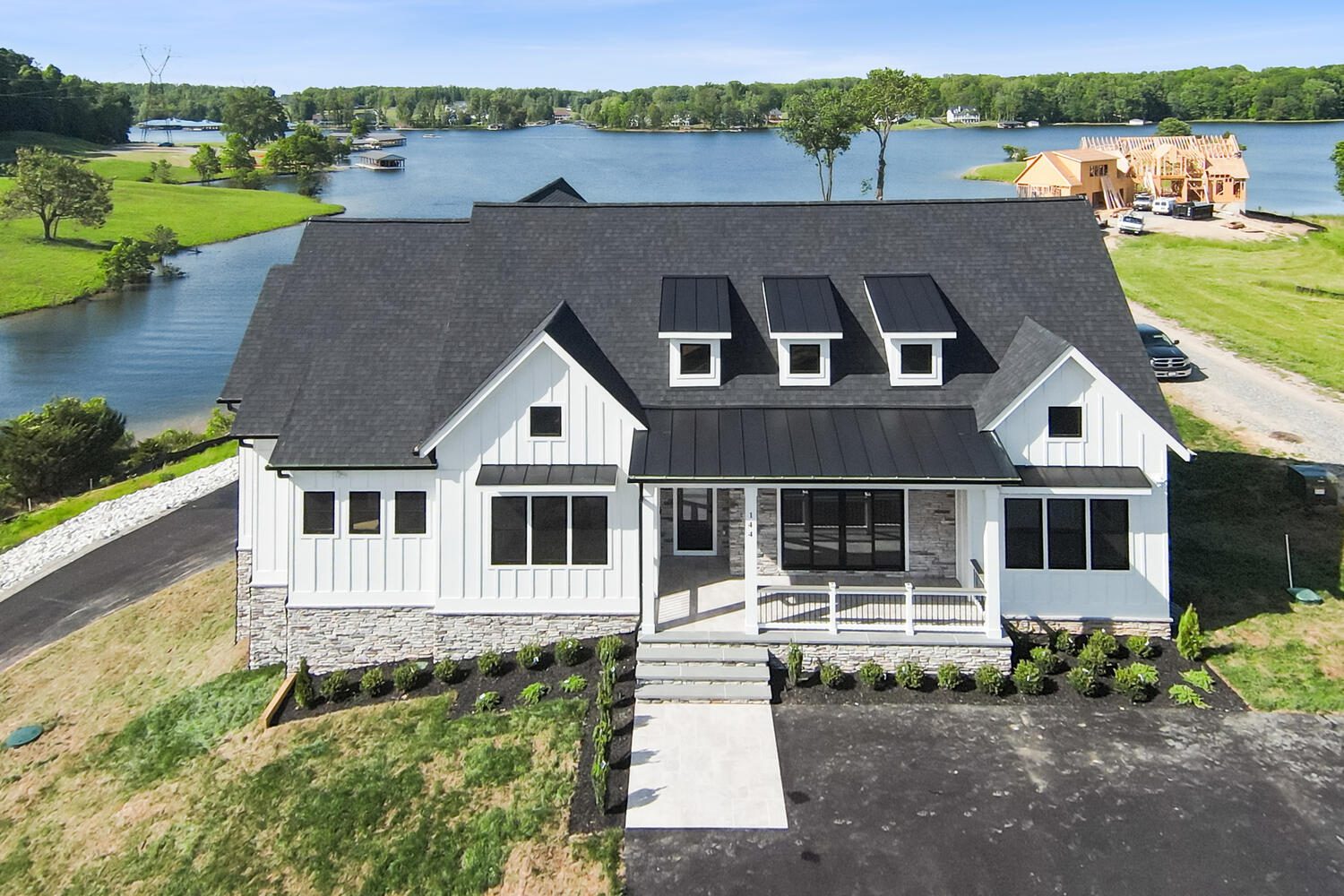 A modern white house with a black roof sits by a lake. It has multiple windows, a small front porch, a driveway, and is surrounded by landscaped grass. Construction is visible nearby.
