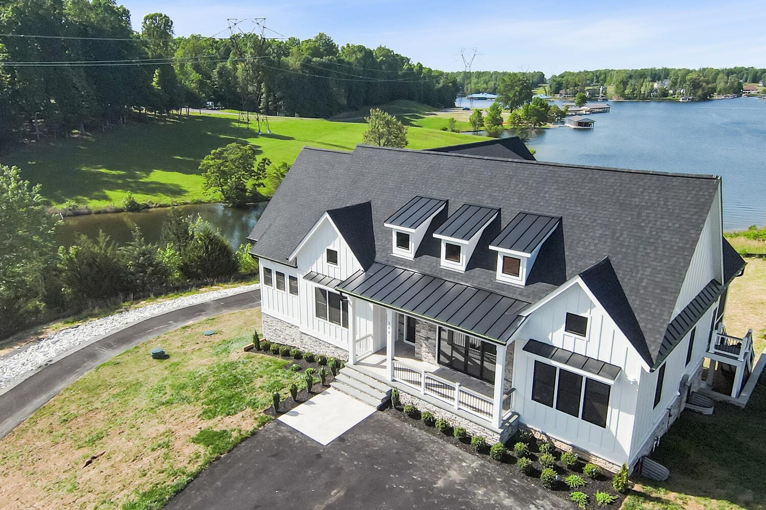 A modern white house with a black roof near a lake. The lawn is partly landscaped, and a driveway leads to the entrance. Trees and water are in the background on a clear day.