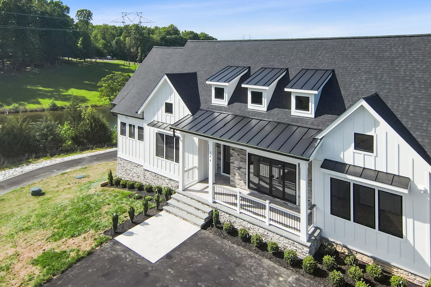 White house with black roof and dormer windows, front porch, and landscaped yard. Located near grassy area and trees under a clear sky.