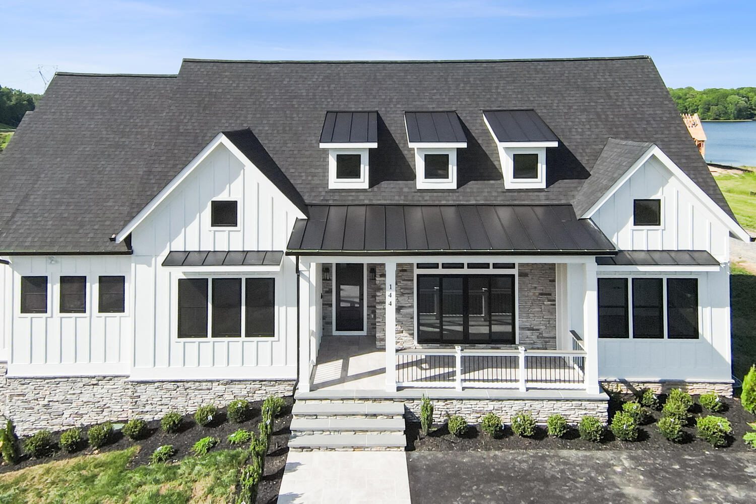 Modern white house with a dark roof, large windows, and a front porch. The facade features stone accents and neatly trimmed bushes. A lake is visible in the background.