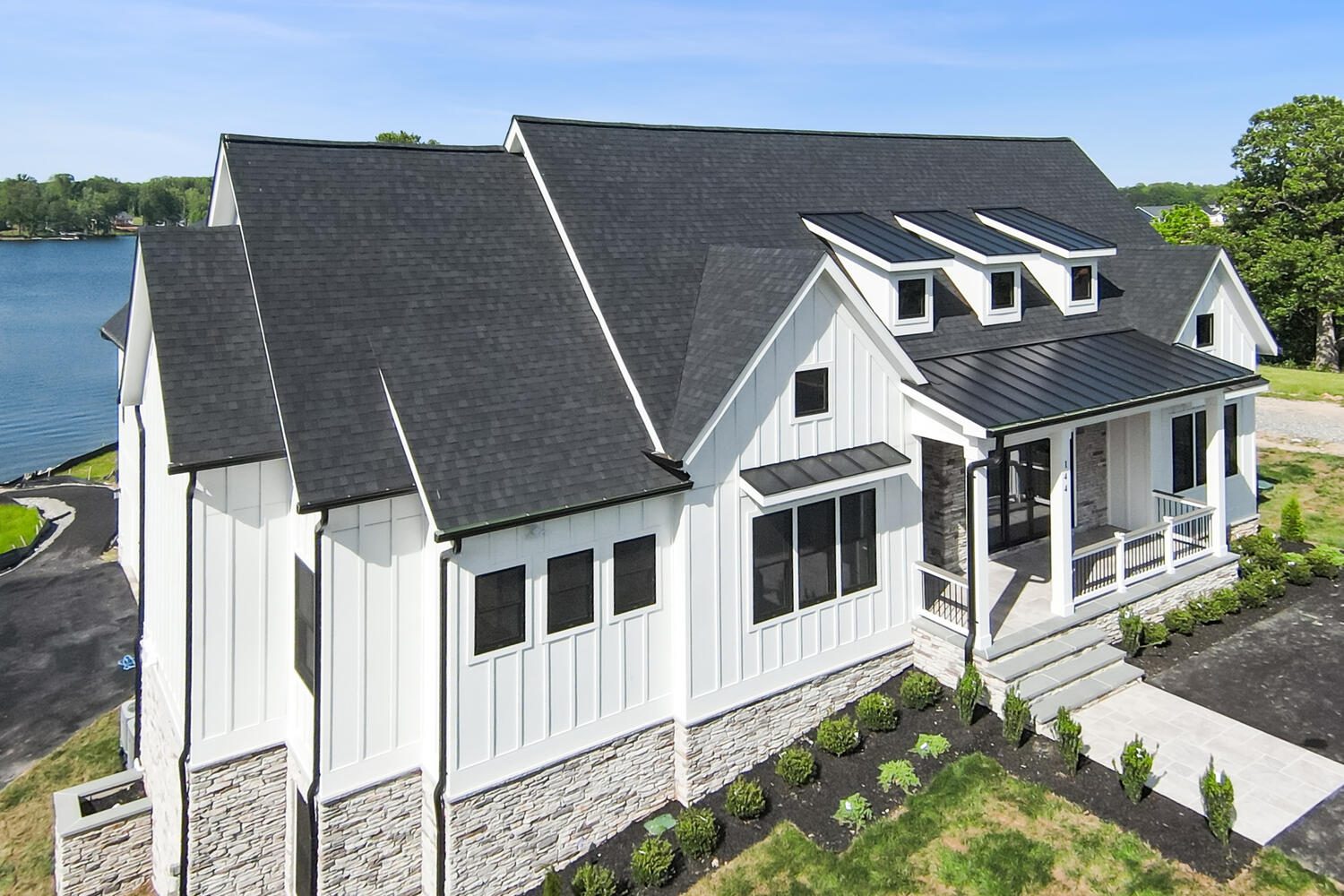 Modern lakefront house with white siding, dark roof, multiple skylights, and a small front porch. The property features a neat, landscaped yard and is situated near a body of water.